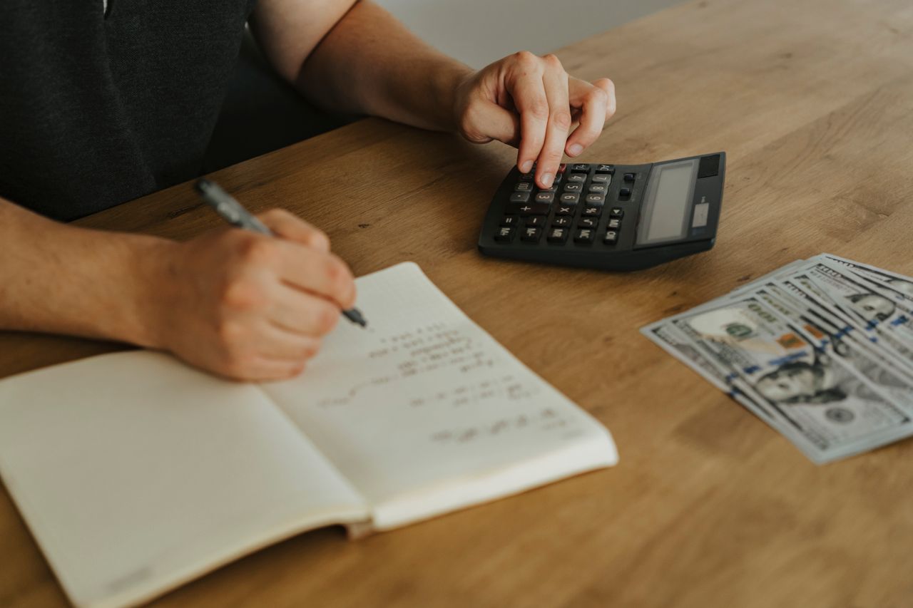 hero-img Close-up of hands using a calculator with notebook and cash on a wooden table.