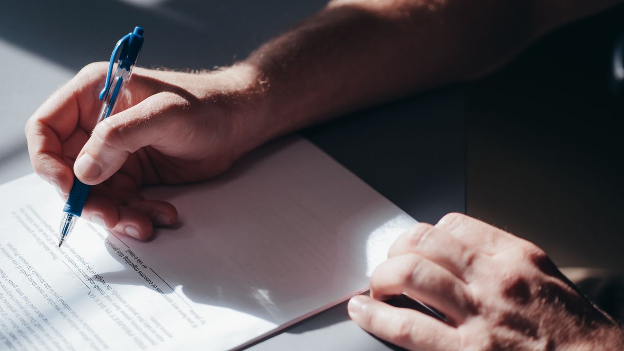who-we-are-img A close-up image of a person's hand signing a document with a blue pen in natural lighting.
